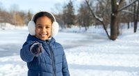 Happy mixed-race boy in blue winter jacket and white earmuffs pointing at camera in snowy park during cold season for outdoor activities and childhood joy.