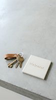 Brass keys on a leather keychain beside a white welcome tile on a concrete surface, symbolizing new home ownership and real estate.
