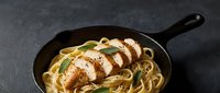 Grilled chicken breast sliced over creamy fettuccine pasta with herbs in cast iron skillet on dark surface for restaurant menu design.