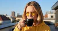 Young blonde Caucasian woman in yellow hoodie drinking coffee on urban rooftop terrace at sunset with city skyline background for lifestyle content.