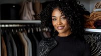 Young African American woman with curly hair smiling in modern walk-in closet with organized clothing racks and luxury fashion items for lifestyle content.
