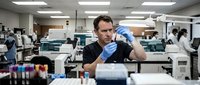 Middle-aged Caucasian male scientist in black shirt examining sample in modern laboratory with colleagues working in background. Research facility setting.