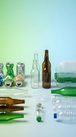 Assorted recyclable materials including glass bottles, plastic containers, aluminum cans and paper products arranged on gradient background for sorting.