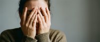 Stressed young woman covering face with hands showing emotional overwhelm and mental health struggles in neutral indoor setting.