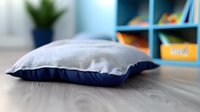 Soft gray and navy blue decorative pillow on wooden surface against blurred modern interior background with bookshelf and plant decor.