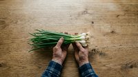 Hands holding fresh green onions with white roots on rustic wooden surface, suitable for cooking, food styling, culinary blogs, and recipe content.