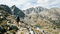 Hiker with backpack standing on rocky mountain trail overlooking alpine lake and jagged peaks, outdoor adventure and wilderness exploration.