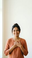 Young South Asian woman with dark hair in casual orange shirt, hands on chest, smiling warmly against neutral background for portrait and lifestyle use.
