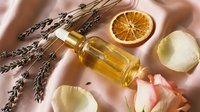 Aromatic serum in glass dropper bottle surrounded by lavender, dried orange slice, and rose petals on soft pink silk fabric.
