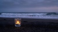 Glowing candle in glass holder on dark beach sand at twilight with ocean waves and purple sky creating peaceful meditation atmosphere.