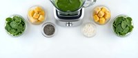 Green smoothie ingredients with spinach, mango, chia seeds and coconut arranged around blender on white background for healthy nutrition recipes.