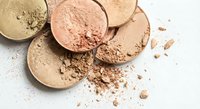 Crushed makeup powder compacts in neutral tones scattered on white surface. Broken cosmetic foundation and blush creating artistic beauty mess for design.