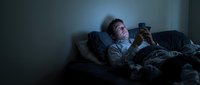 Young Caucasian man lying in bed using smartphone in dark bedroom with blue screen light illuminating face during late night hours.