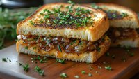 Grilled sandwich with caramelized onions and fresh herbs on toasted bread, served on wooden cutting board. Close-up view shows melted filling and garnish.