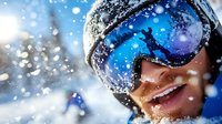 Smiling skier wearing protective goggles during snowfall on winter mountain slope, close up portrait against bright snowy background with bokeh effect.
