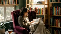 Young woman reading book in cozy home library with bookshelves, sitting in comfortable armchair by window. Relaxing indoor lifestyle scene.