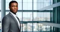 Young African American male professional in gray business suit and tie standing in modern office building with floor-to-ceiling windows overlooking cityscape, suitable for corporate, business, and leadership concepts.