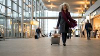 Middle-aged Caucasian businesswoman walking through modern airport terminal with rolling suitcase, wearing burgundy coat and professional attire.