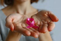 Female cupped hands gently holding small pink awareness ribbon against soft neutral background, symbolizing support for breast cancer awareness campaign.
