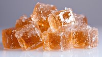 Brown sugar crystals in cube shapes with rough texture and sparkling surface against light gray background, macro food photography showing sweet ingredient details.