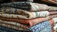 Stack of folded vintage floral pattern textiles in blue, coral and cream colors showing delicate ornamental designs and traditional fabric prints.