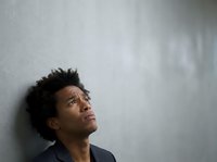 Thoughtful African American man with short dark hair wearing dark blazer looking upward against neutral gray wall background, expressing contemplation and introspection.