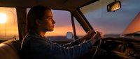 Young woman driving vintage car during golden hour sunset with warm atmospheric lighting creating nostalgic road trip adventure mood.