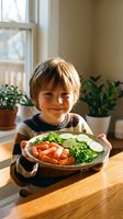 Happy young boy holding plate of fresh vegetables including carrots, cucumbers and greens in bright kitchen with natural light and houseplants.