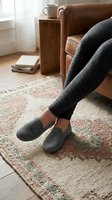 Woman relaxing at home in gray knit slippers and leggings, sitting on leather armchair on a cozy patterned rug.