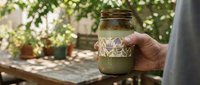 Hand holding a decorative green glass jar with floral pattern in a sunlit garden setting with potted plants in the background.