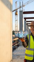 Male construction worker in safety gear holding digital communication icons at building site with cranes and steel framework.