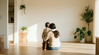 Young mother and toddler son sitting together on wooden floor in bright minimalist living room with houseplants creating warm family bonding moment at home.