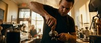 Focused male barista in black apron tamping espresso grounds in a busy cafe, warm light and steam creating an authentic coffee shop atmosphere.