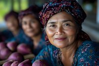 Asian senior woman vendor in traditional headscarf smiling while selling fresh purple mangoes at local market, authentic cultural portrait showing joy and contentment.