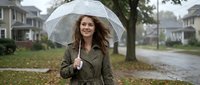 Young woman with long dark hair and fair complexion holding transparent umbrella in residential neighborhood during rainy autumn day, wearing olive jacket for weather protection.