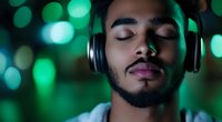 Young African American man with closed eyes and beard wearing headphones, experiencing peaceful moment with green bokeh lights background. Music meditation concept.
