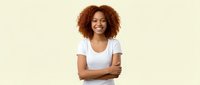 Happy young African American woman with curly hair smiling confidently with arms crossed wearing white t-shirt on beige background for lifestyle content.