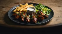 Grilled chicken wings with barbecue glaze served on black plate with golden french fries, fresh salad and creamy dipping sauce on rustic wooden table.