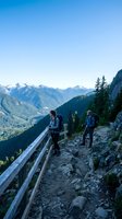Two hikers with backpacks walking on mountain trail with wooden railings overlooking alpine valley and snow-capped peaks under clear blue sky.