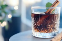 Crystal glass with dark cocktail garnished with cinnamon stick and mint leaf on marble surface, soft bokeh lights background creates cozy atmosphere.