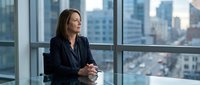 Professional businesswoman in dark suit sitting at conference table in modern office with city skyline view through large windows for corporate presentations.