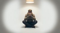Young man sitting cross-legged on floor holding head in hands showing stress, depression, mental health struggles in minimalist white room with overhead lighting.