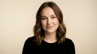 Young professional Caucasian woman with wavy brown hair smiling confidently in black top against neutral beige background for business portraits.