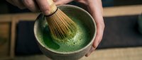 Hands whisking matcha powder in ceramic bowl creating frothy green tea for traditional Japanese tea ceremony preparation and wellness beverages.