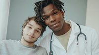 Young African American male doctor with dreadlocks and stethoscope posing with Caucasian male patient in medical office setting for healthcare consultation.
