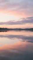 Serene lake at sunset with soft pink and purple sky reflecting on calm water, forested shoreline in distance, peaceful nature landscape for relaxation and wellness projects.