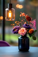 Elegant floral arrangement with purple anemones and coral roses in black textured vase, illuminated by vintage pendant lamp with warm bokeh background.