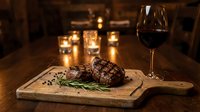 Grilled beef steak with herbs on wooden board paired with red wine glass in cozy restaurant setting with warm candlelight ambiance for dining experience.