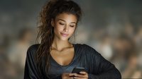 Young mixed-race woman with curly hair looking at smartphone, wearing casual dark clothing against blurred background. Soft lighting creates intimate atmosphere.