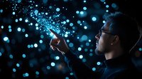 Asian man in glasses pointing at glowing blue particles, side view against dark background with bokeh lights creating magical atmosphere for technology concept.